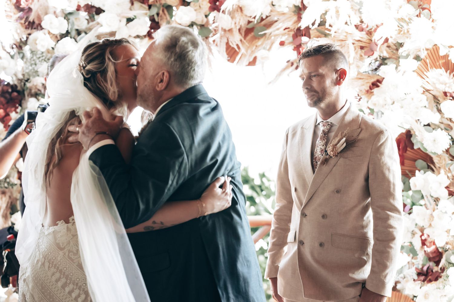A bride shares a kiss with an older man at the altar while a groom in a beige suit watches at a floral wedding.