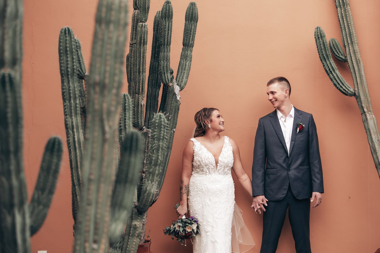 A bride and groom hold hands and smile at each other beside tall saguaro cacti against a terracotta wall.