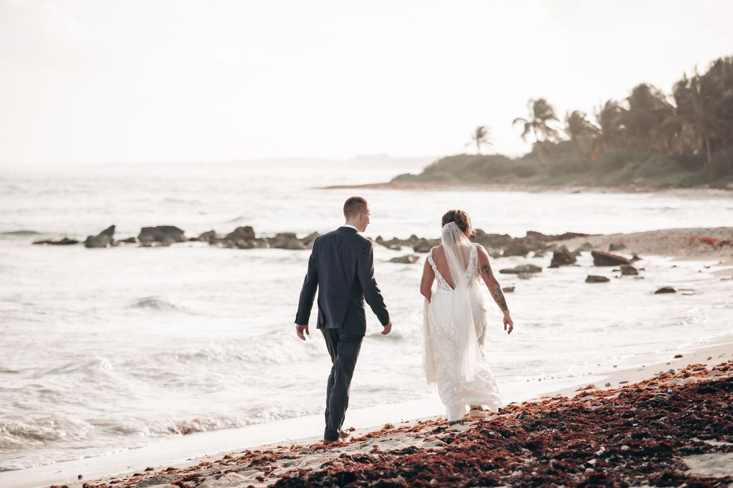 Bride and groom walking hand in hand along a rocky tropical beach at sunset.