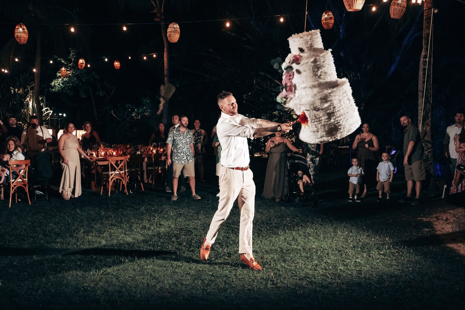 Groom swings at a wedding cake-shaped pinata at an outdoor evening reception.