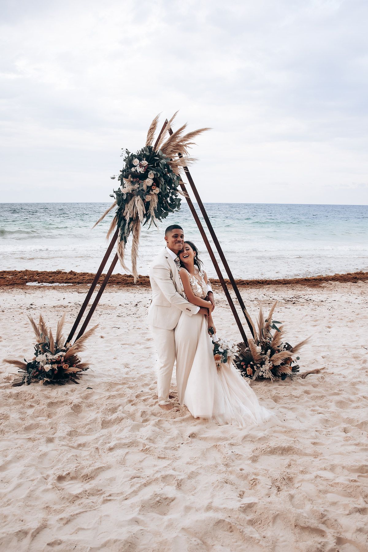 Beachside wedding ceremony with wooden triangle arch and tropical floral decorations on sandy shore.