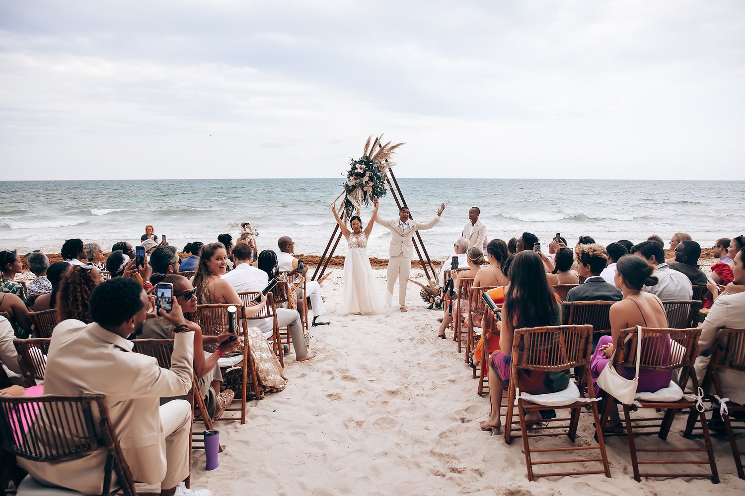 Beach wedding ceremony setup with wooden chairs arranged in rows facing the ocean under a bright sky.