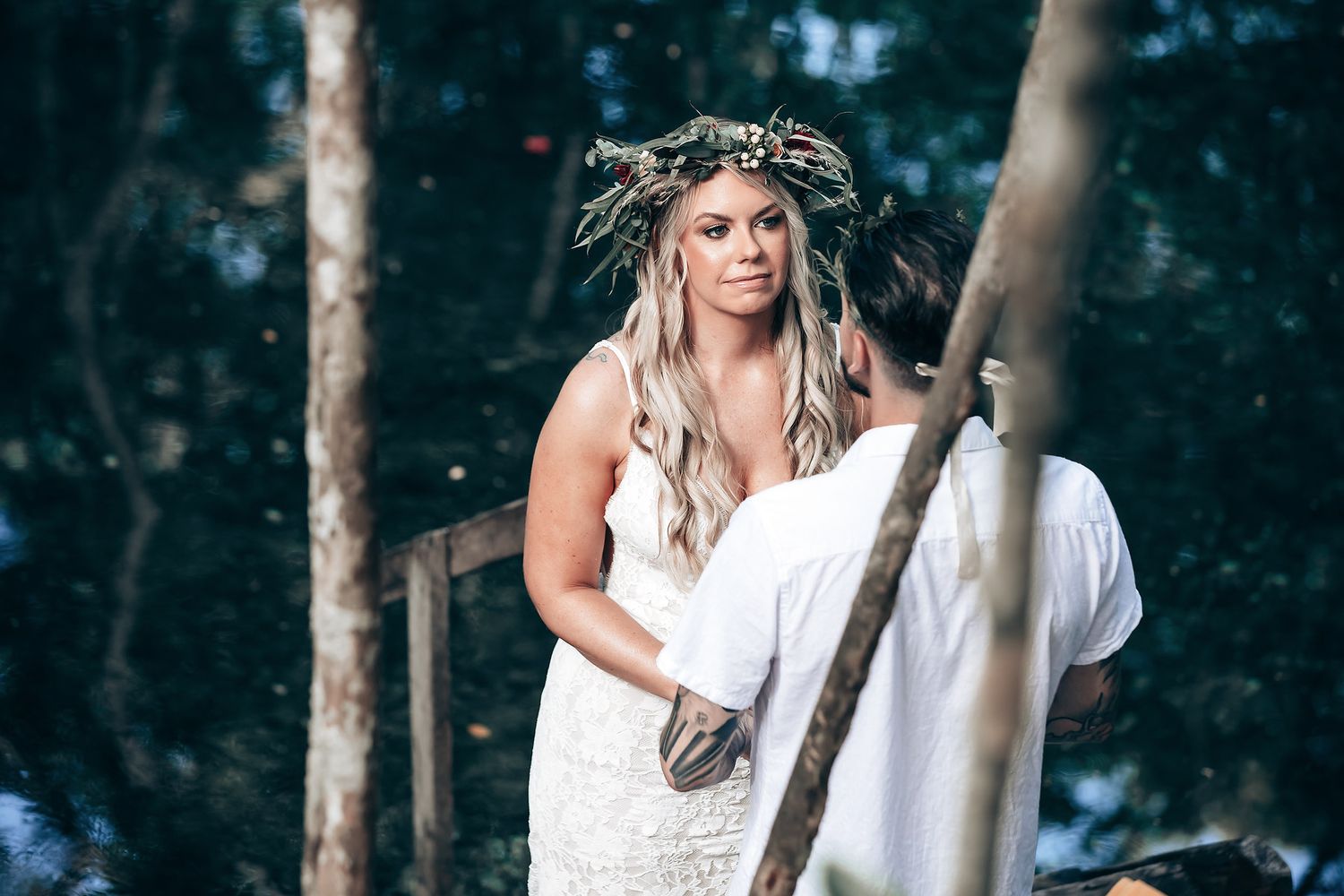 Bride in lace dress and floral crown stands in mystical forest setting during ceremony.