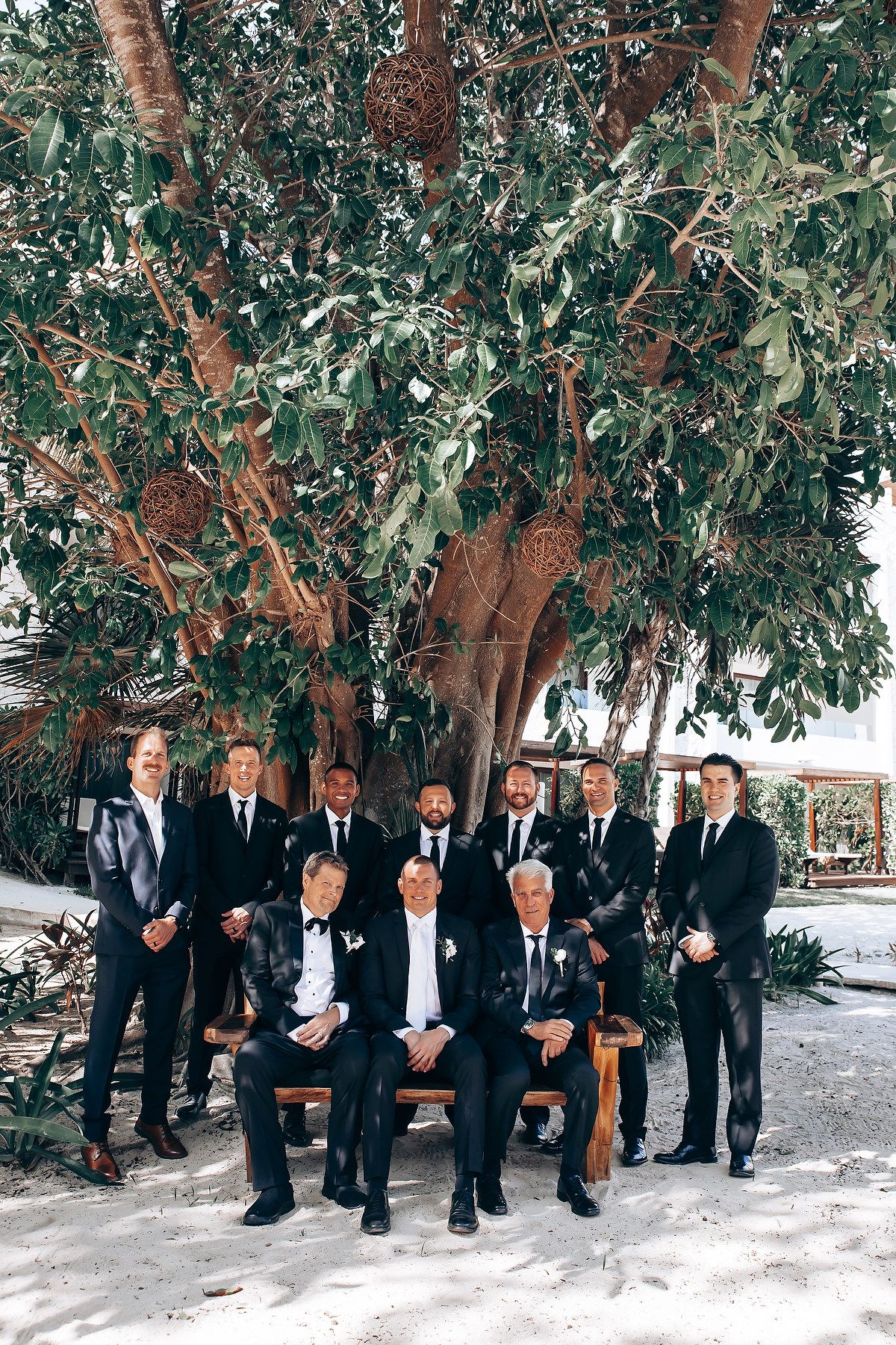 Groomsmen in black suits gather under a shaded tree area for wedding party photos.