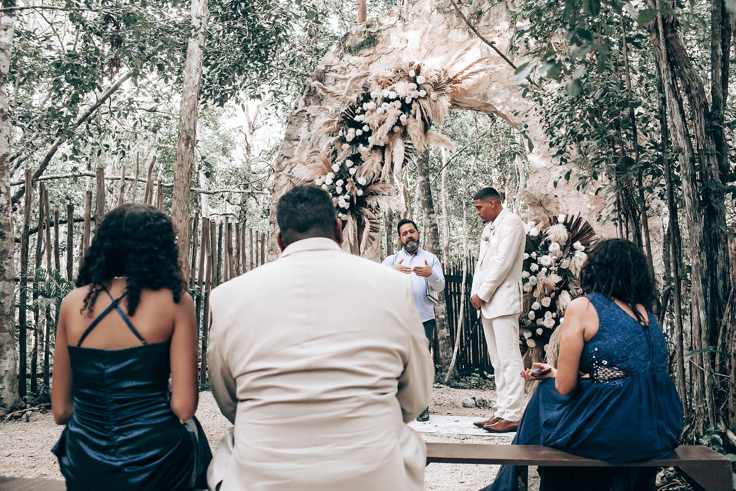Wedding ceremony guests seated outdoors among trees in formal attire.