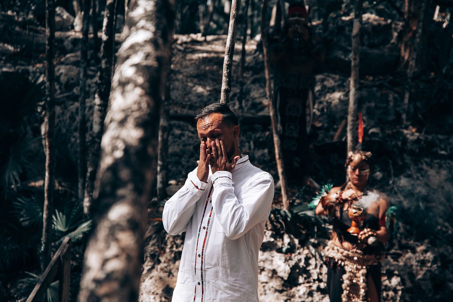 People in white clothing stand among trees in a dark, atmospheric forest setting.