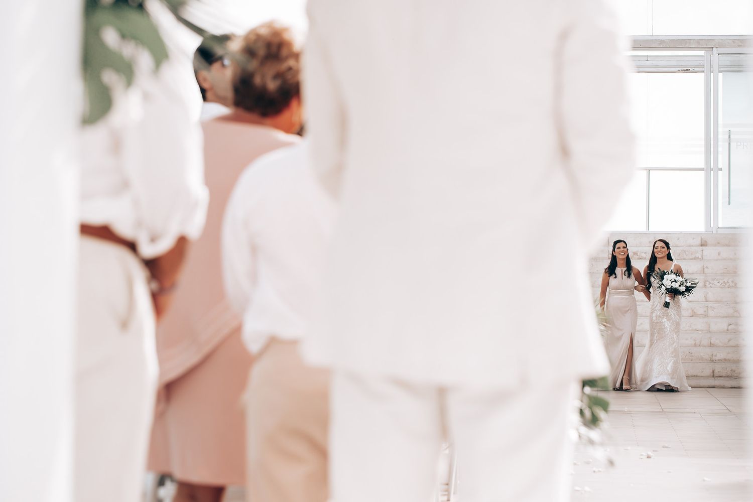 Wedding party in white attire stands in a bright hallway while bride and bridesmaid are seen in the distance.
