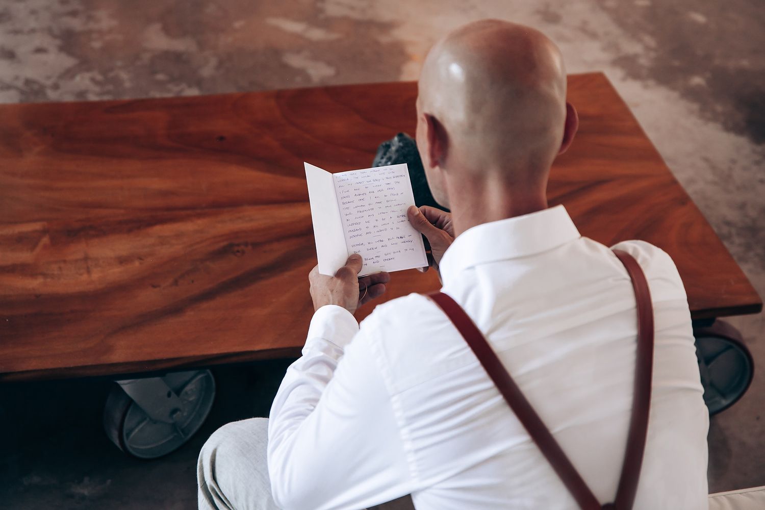 A person in a white shirt and suspenders sits at a wooden table reading a paper document.
