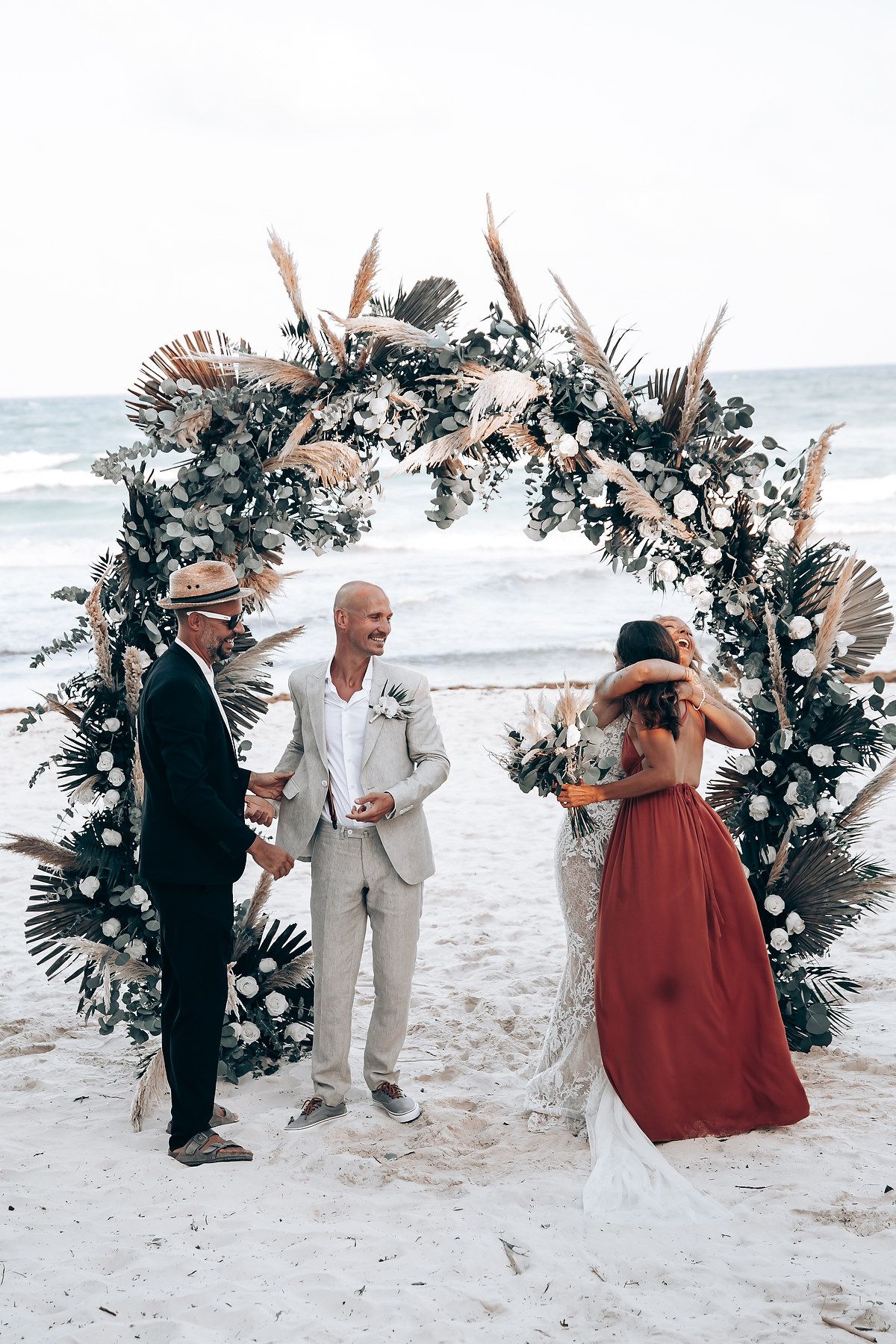 Beach wedding ceremony with large floral arch and guests gathered on sandy shore.