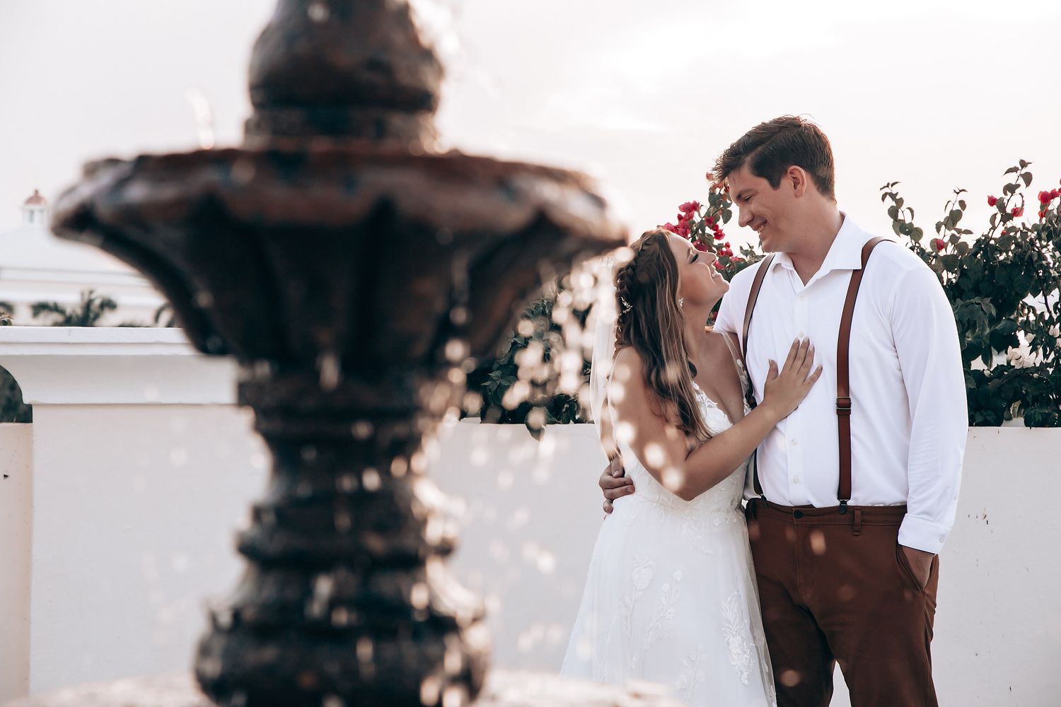 Romantic couple in wedding attire share an intimate moment next to an ornate fountain at sunset.