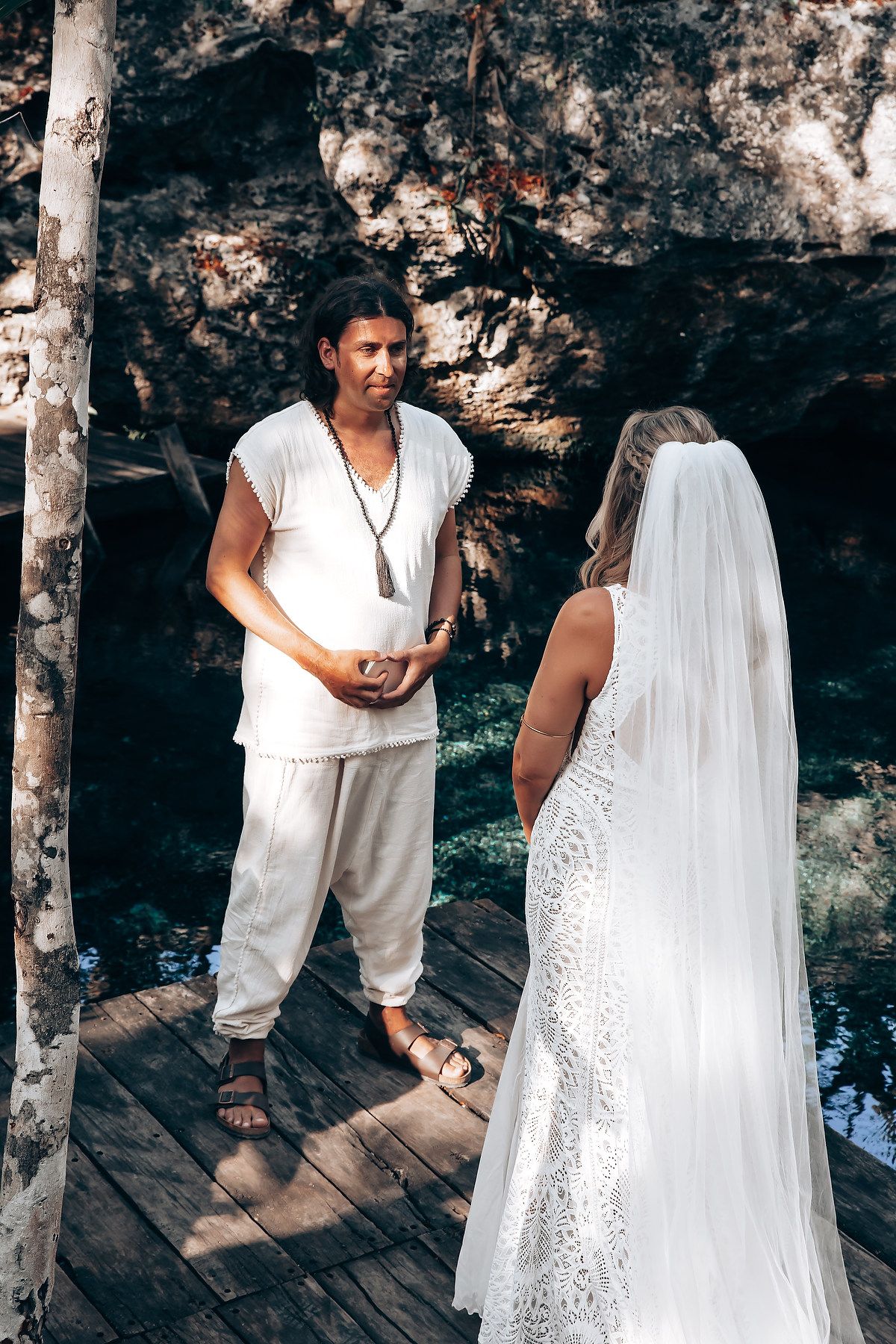 A couple in white attire stands together on a wooden deck surrounded by trees in a romantic outdoor setting.