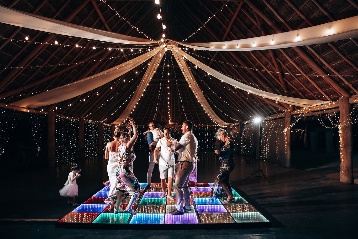 Illuminated LED dance floor panels glow under draped string lights at an indoor wedding reception venue.