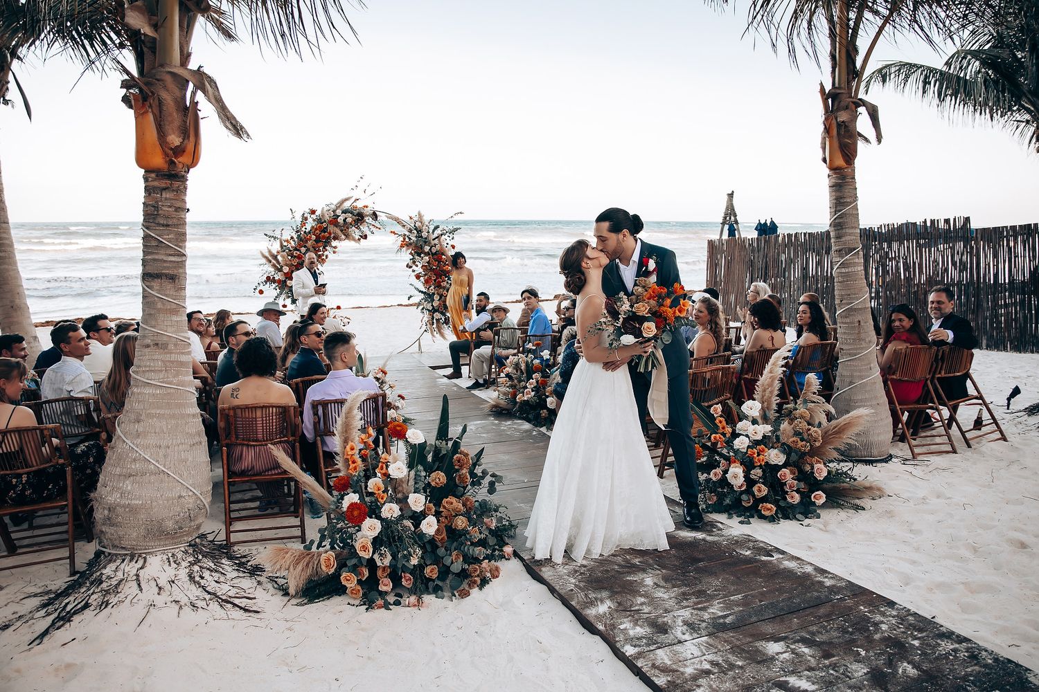 Wedding guests gather on a tropical beach with palm trees and ocean views during a sunset ceremony.