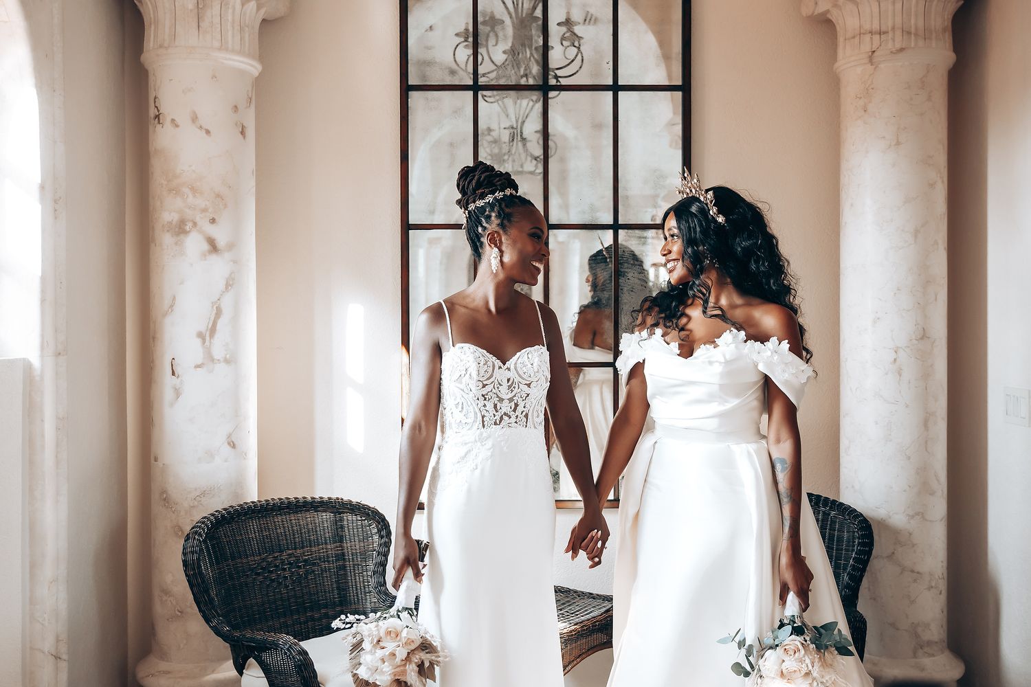 Two people in elegant white wedding gowns hold hands in a sunlit venue.