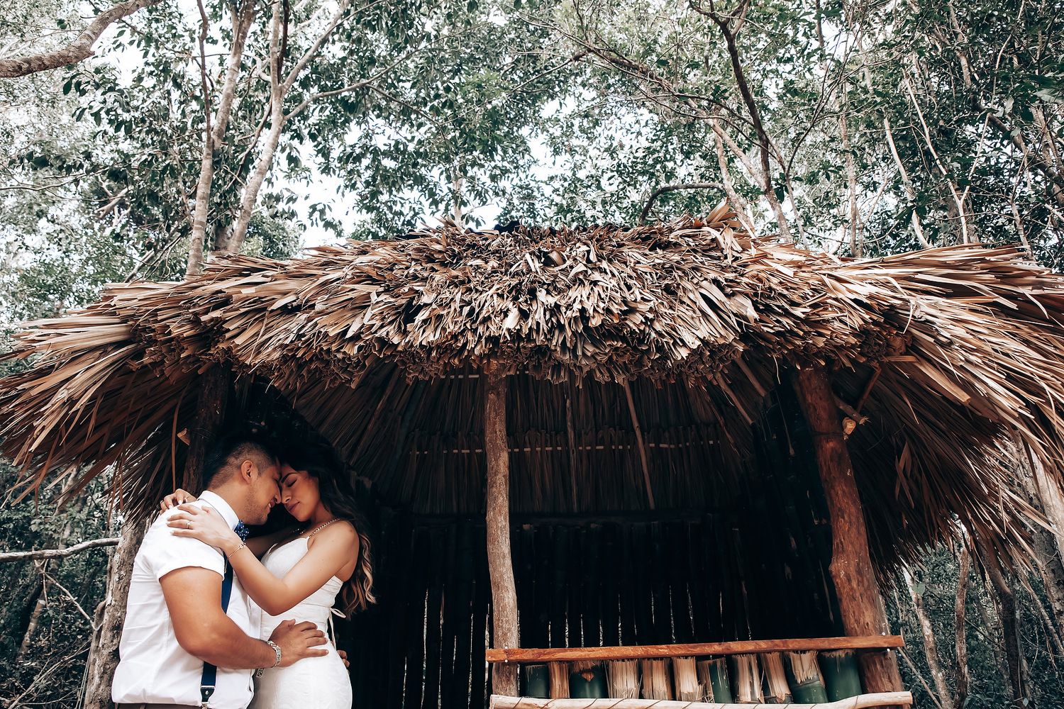 A couple embraces in front of a thatched-roof hut surrounded by tropical foliage and natural scenery.