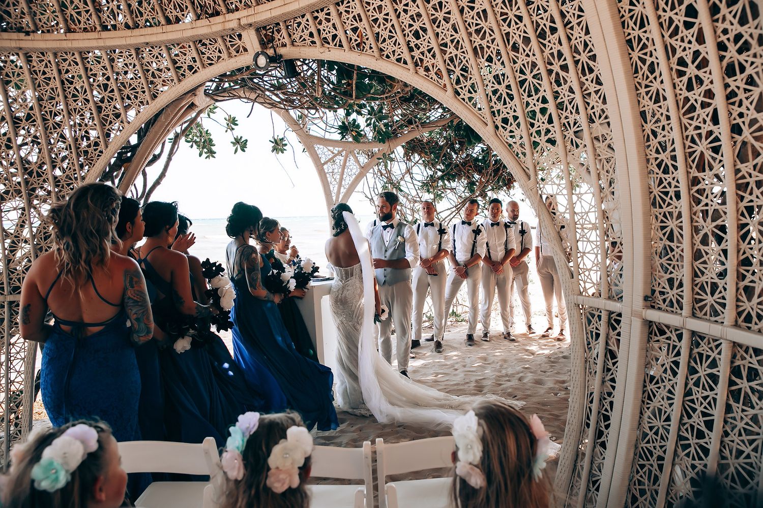 Wedding guests gather under an ornate latticed canopy structure during an outdoor ceremony.