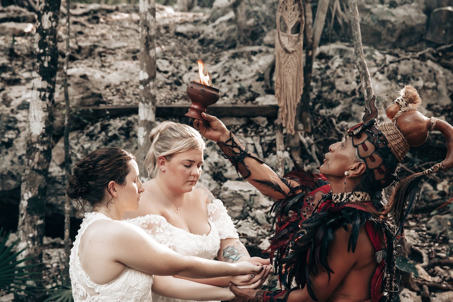 Two brides in white wedding dresses embrace a tribal man in a lush, outdoor setting.