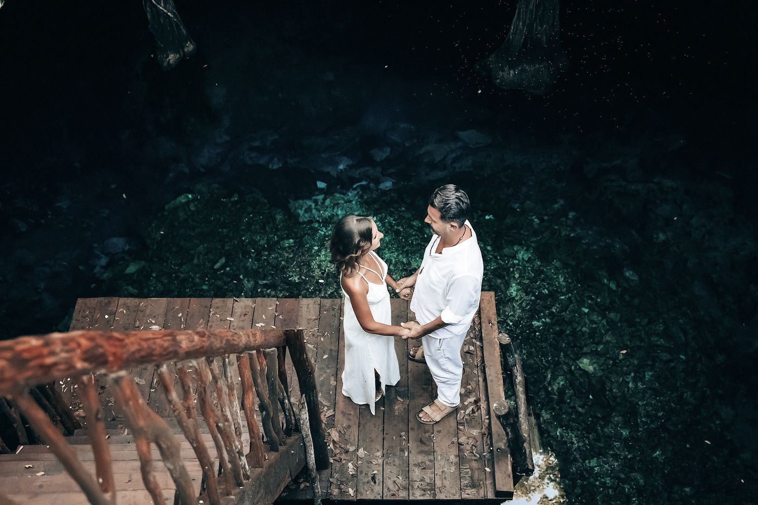 Two people in white clothing standing on a wooden bridge over a body of water.