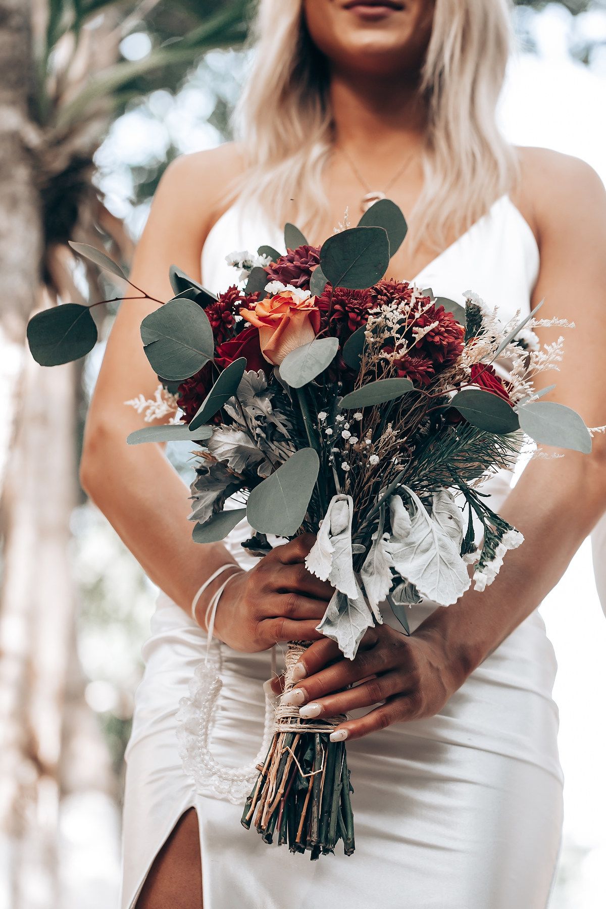 A wedding bouquet with burgundy flowers, white blooms and eucalyptus held against a white bridal gown.
