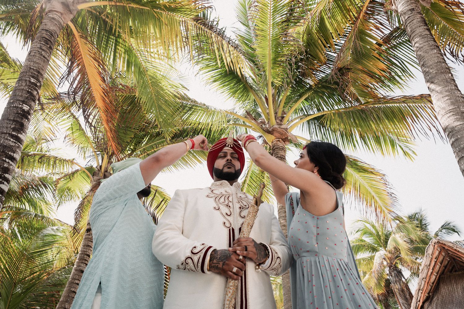Sikh groom in red turban and white sherwani during outdoor Indian wedding ceremony under palm trees.
