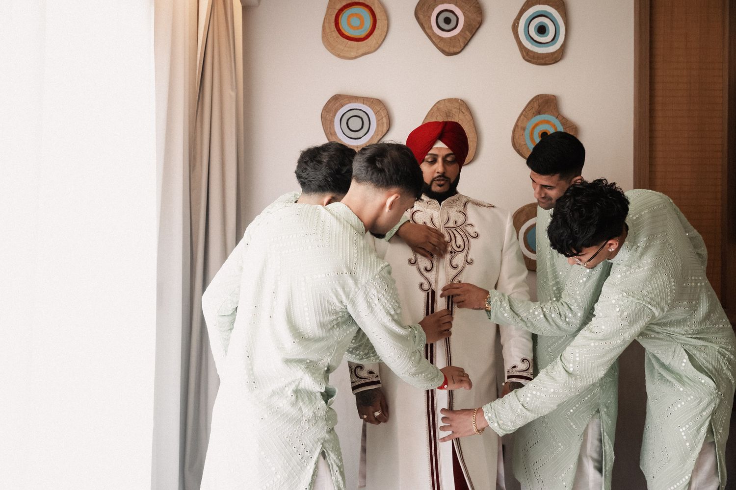 Sikh groom in red turban and white sherwani being dressed by groomsmen in matching outfits before wedding ceremony.