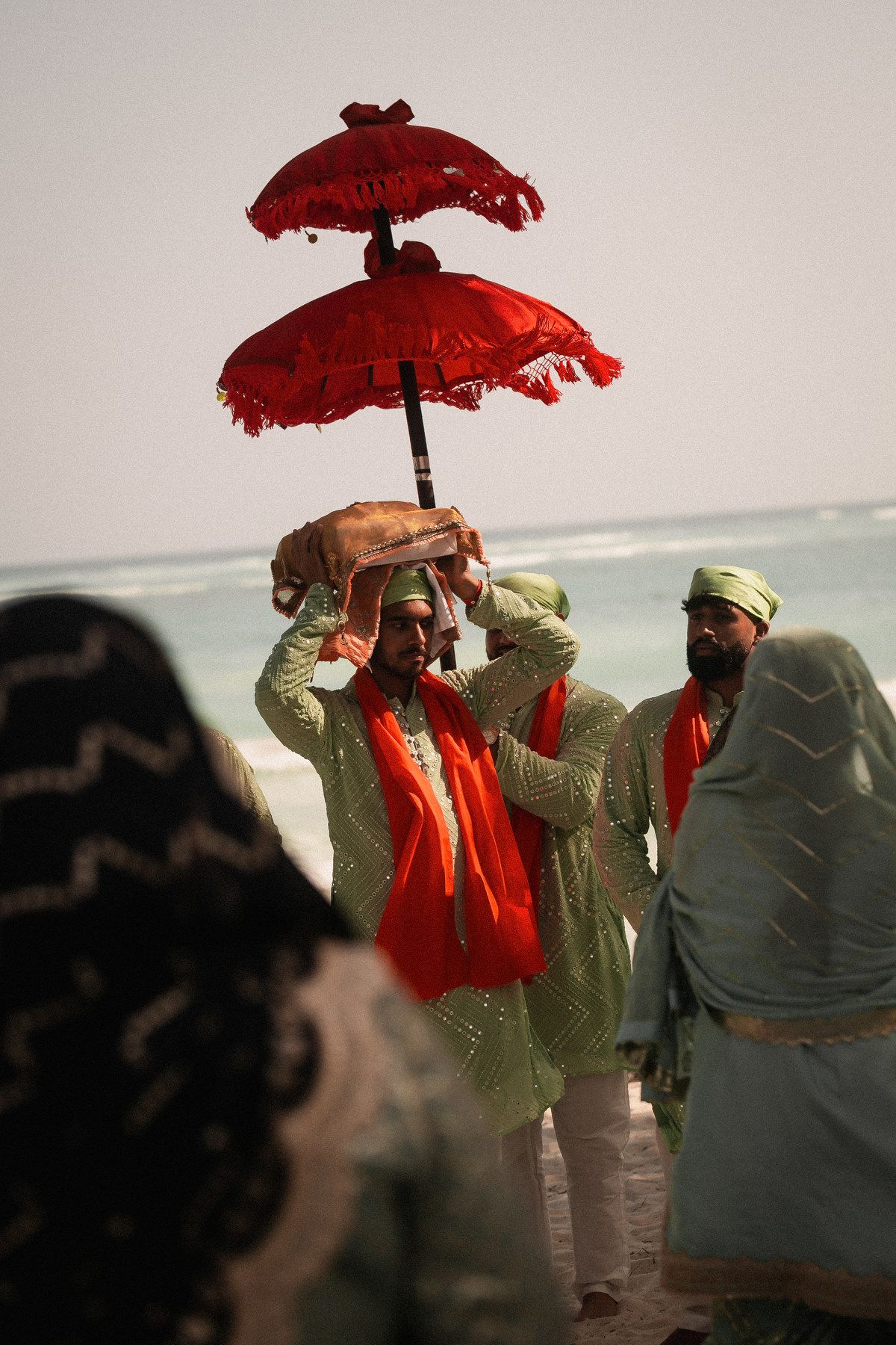 Man carries holy Guru Granth Sahib under red ceremonial umbrella on beach at Sikh wedding.