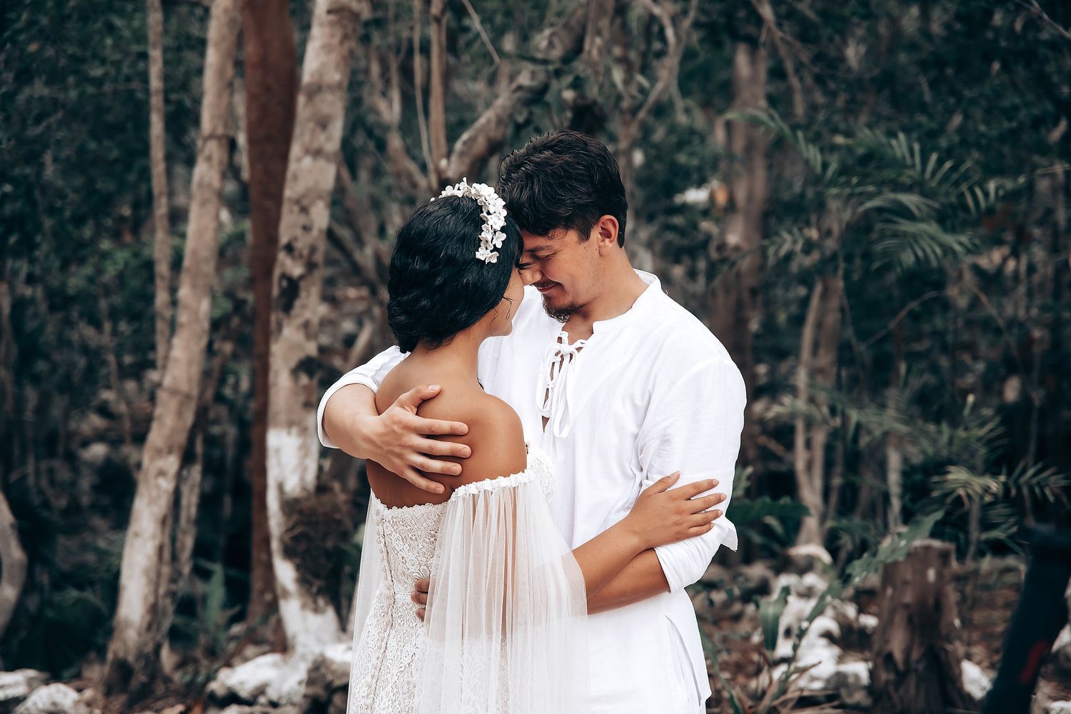 A couple embraces tenderly on a wooden boardwalk with lush forest greenery in the background.