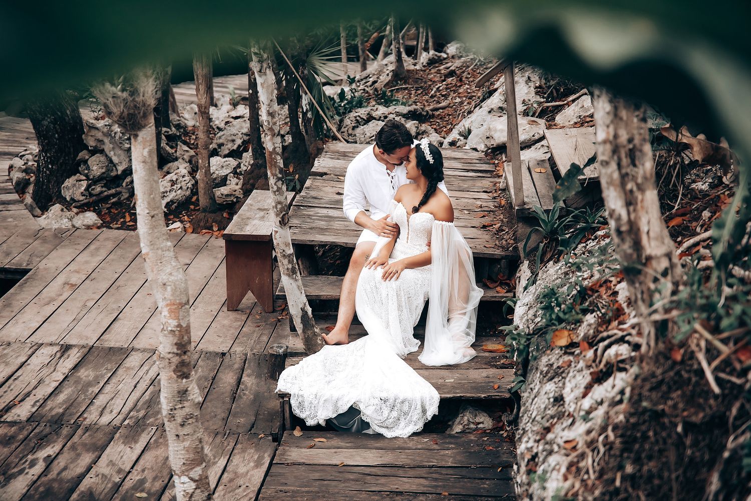 A person in a white dress sits gracefully on rustic wooden steps surrounded by natural foliage and dappled sunlight.