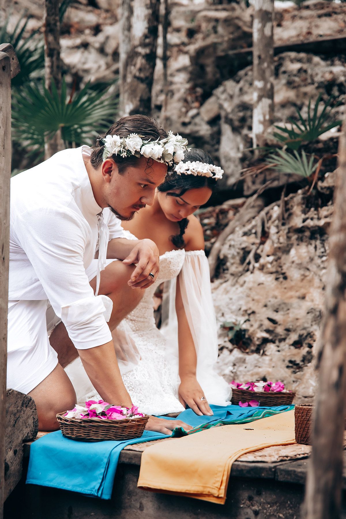 Two people in white attire lean over a table with colorful fabrics and baskets in an outdoor setting.