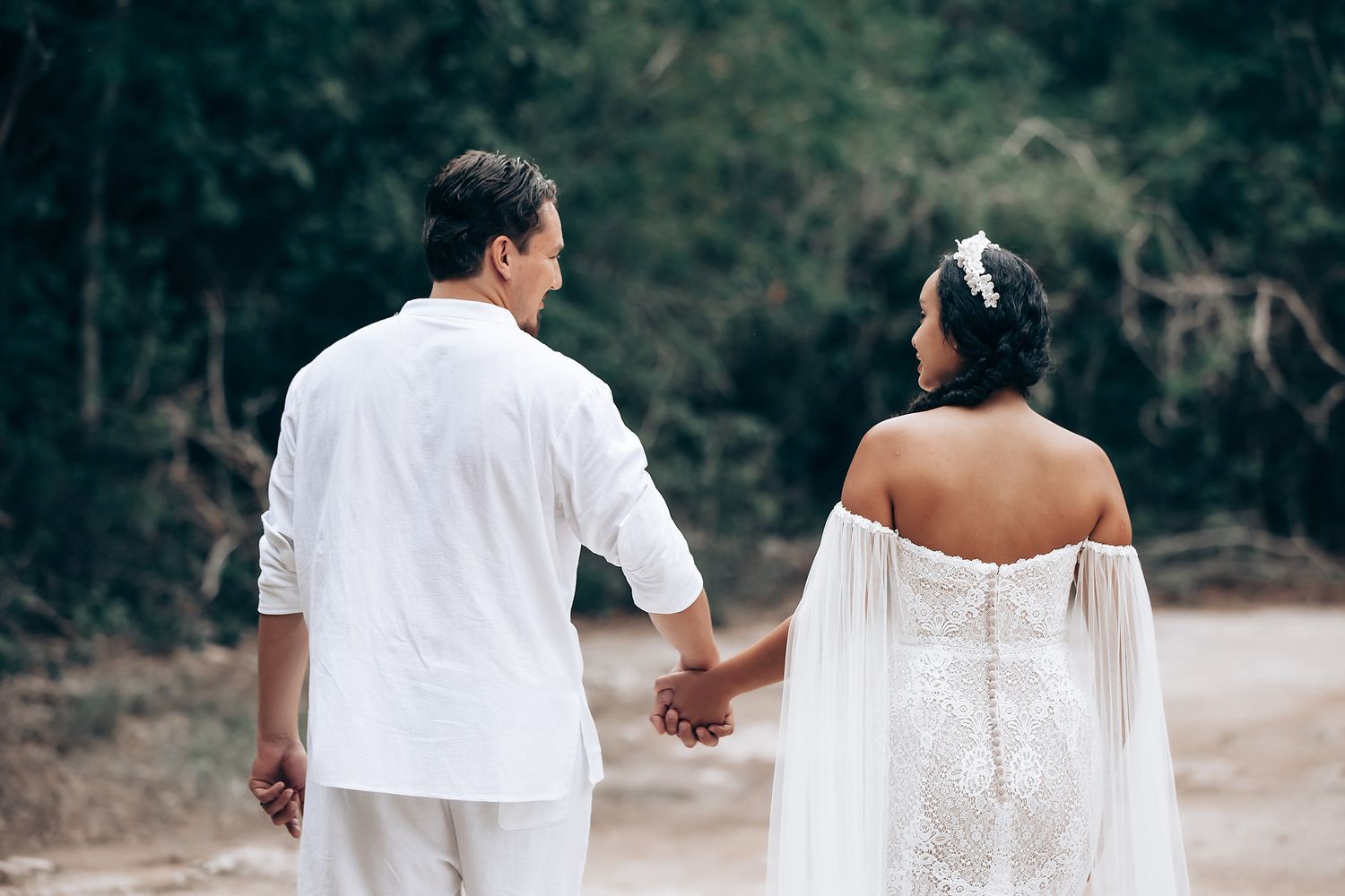 A couple in white wedding attire walking hand in hand through a scenic outdoor setting.