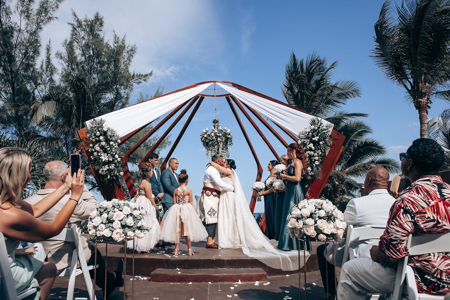 Wide angle view of beach wedding ceremony with guests and palm trees against blue sky.