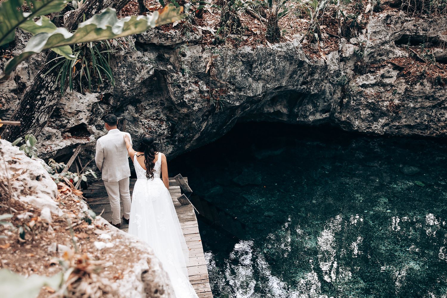 Couple in wedding attire walks along rocky coastal cliffs near dark ocean caves and rugged terrain.