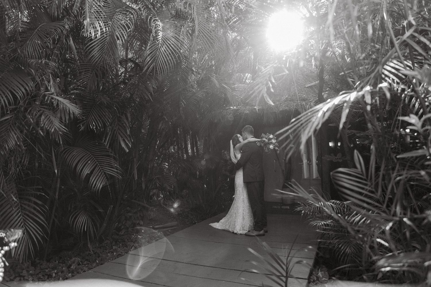 Bride and groom share romantic first kiss surrounded by lush tropical greenery in black and white wedding photo.