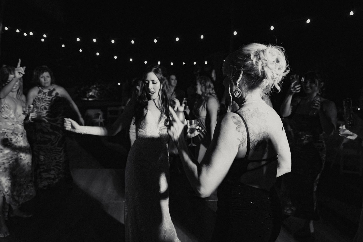 Black and white photo of bride and wedding guests dancing joyfully under string lights at reception.
