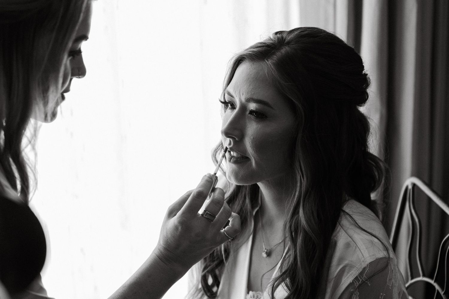 Black and white photo of bridesmaid applying makeup to bride before wedding ceremony.