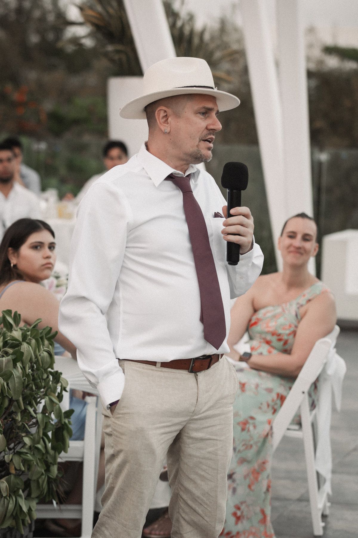 Man in white shirt, burgundy tie, and panama hat gives a speech holding microphone at wedding reception.