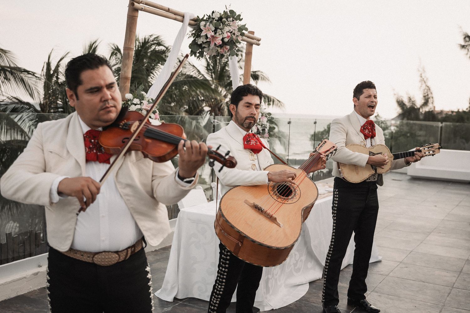 Mariachi band playing violin and guitar at an outdoor wedding ceremony with floral arch backdrop.