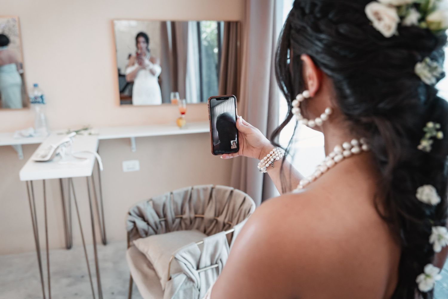 Bride in pearl necklace taking selfie in mirror wearing lace wedding gown with floral hair accessories.
