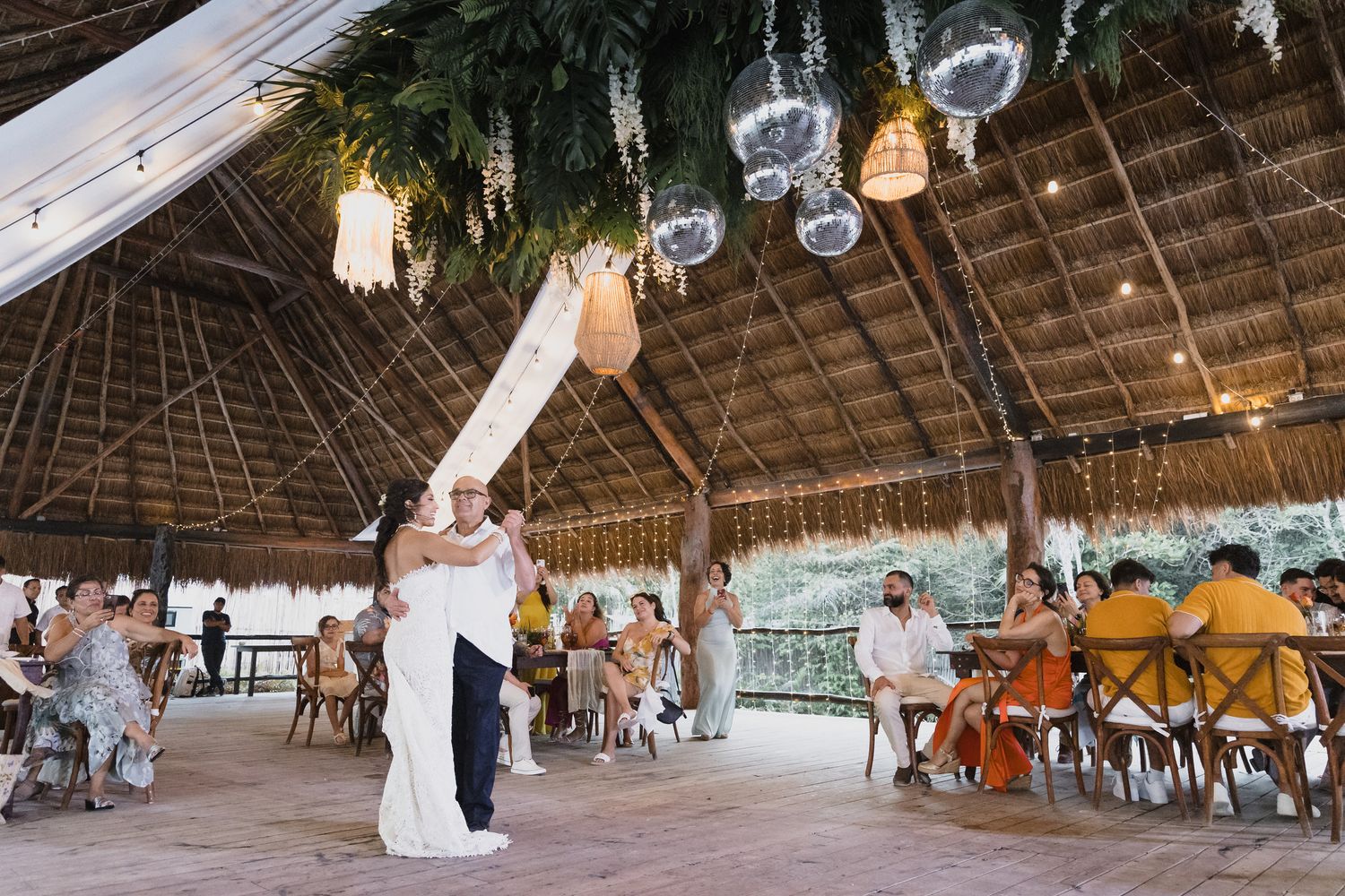 Bride and groom share first dance under palm-thatched pavilion with string lights.