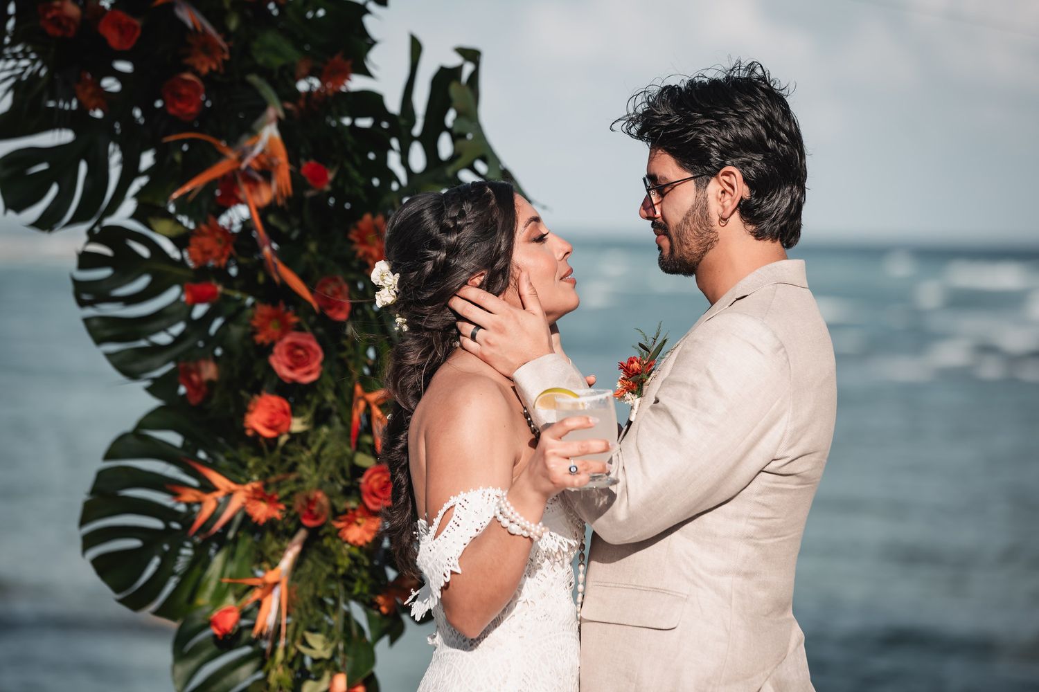 Bride and groom share romantic moment under tropical floral arch with ocean in background.