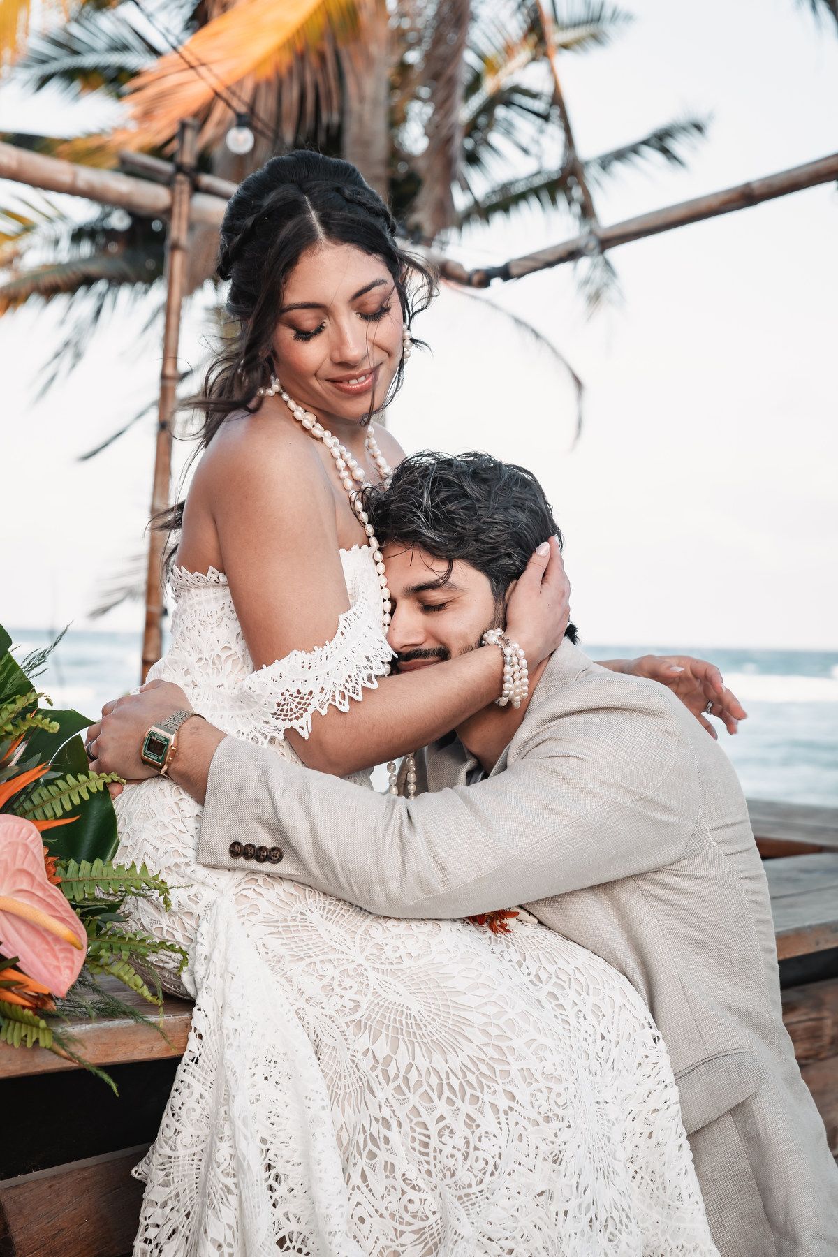 Bride in lace gown tenderly embraces groom at sunset tropical beach wedding reception.
