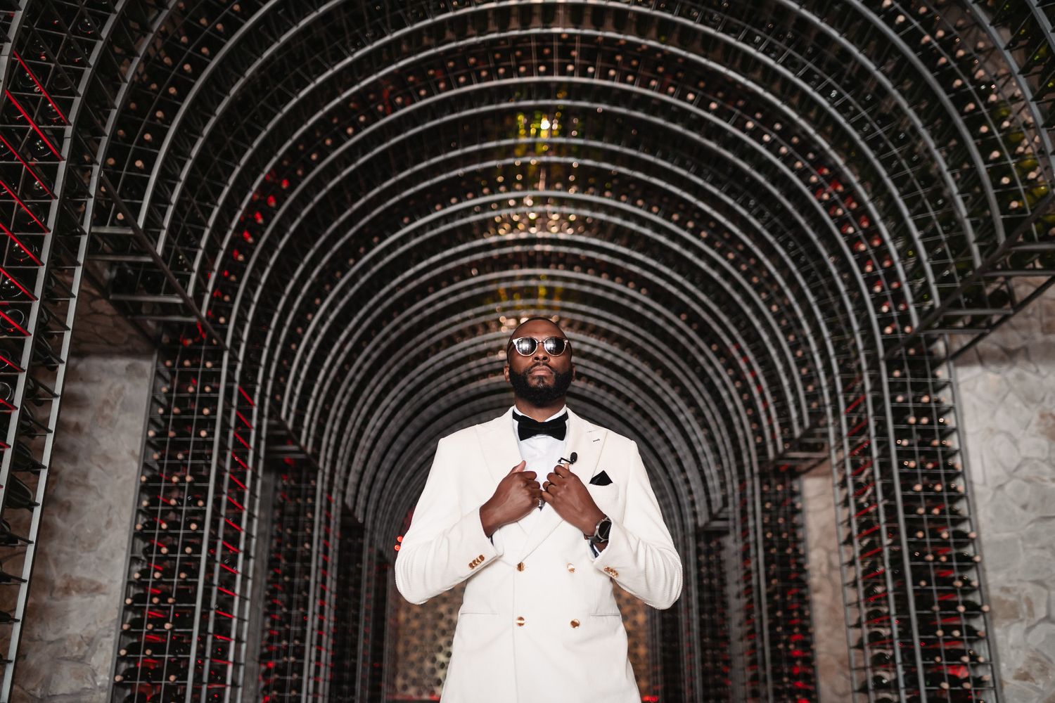 Man in white tuxedo poses in elegant wine cellar with arched bottle-lined walls.