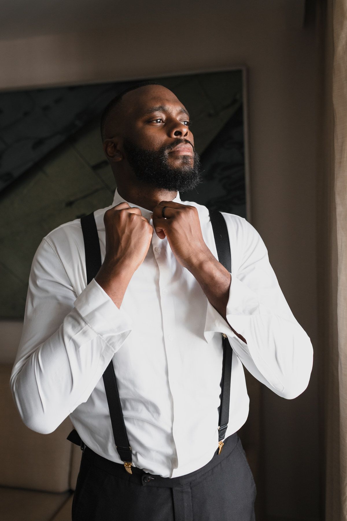 Dapper groom in white shirt with suspenders adjusting bow tie in hotel room.