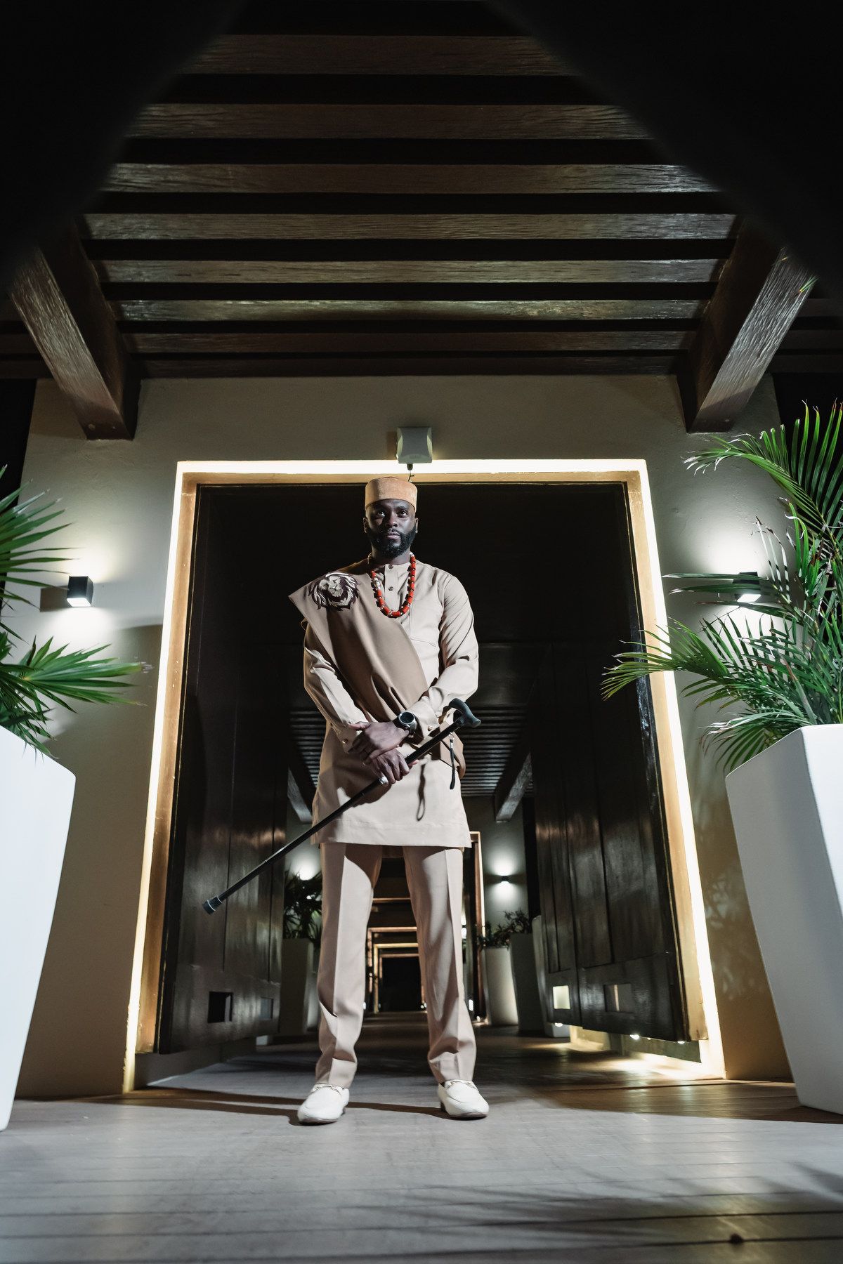Man in traditional African attire holding a ceremonial staff standing in illuminated doorway.