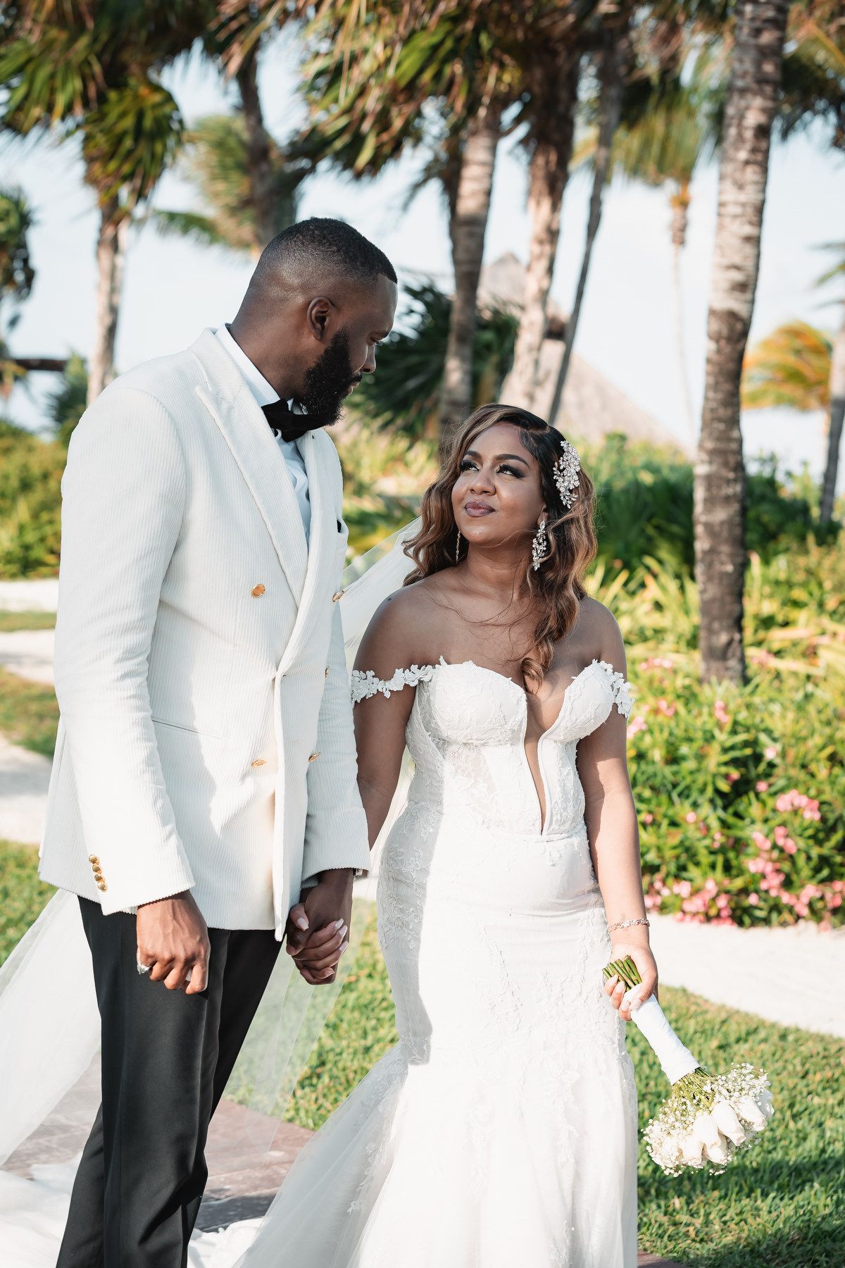 Interracial wedding couple holding hands outdoors among tropical palm trees at a destination wedding.