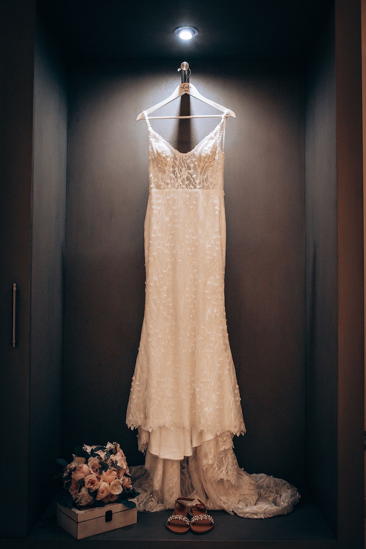 Elegant white lace wedding dress displayed on a hanger against a dark background with soft lighting.