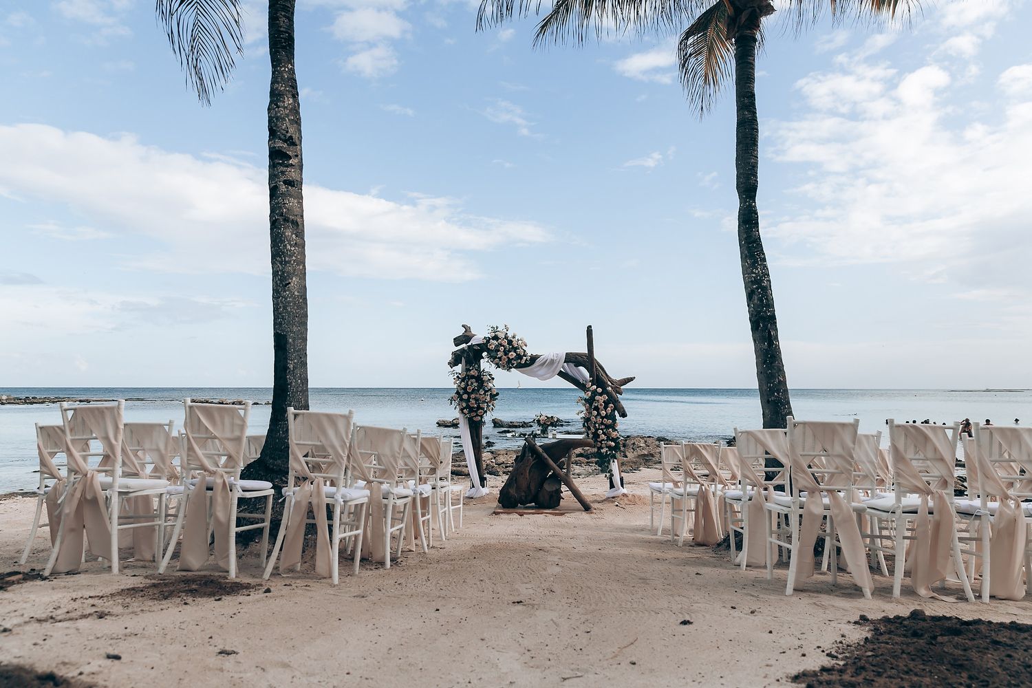 Beach wedding setup with white chairs and palm trees overlooking the ocean.