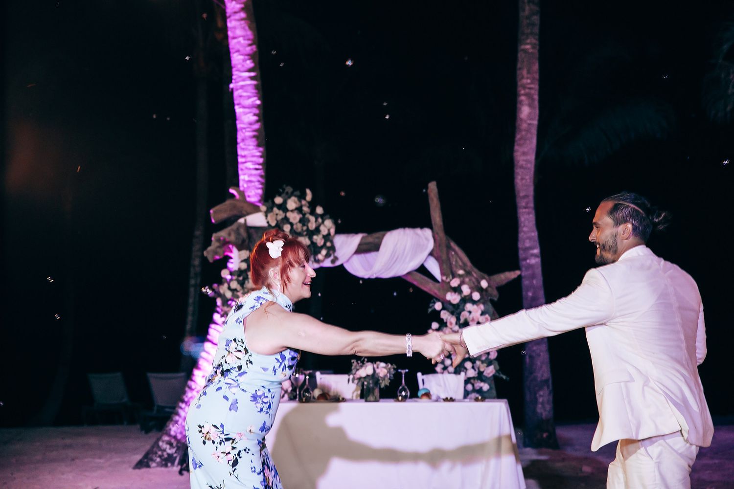 Wedding party dancing and celebrating during a nighttime beach reception with purple accent lighting.