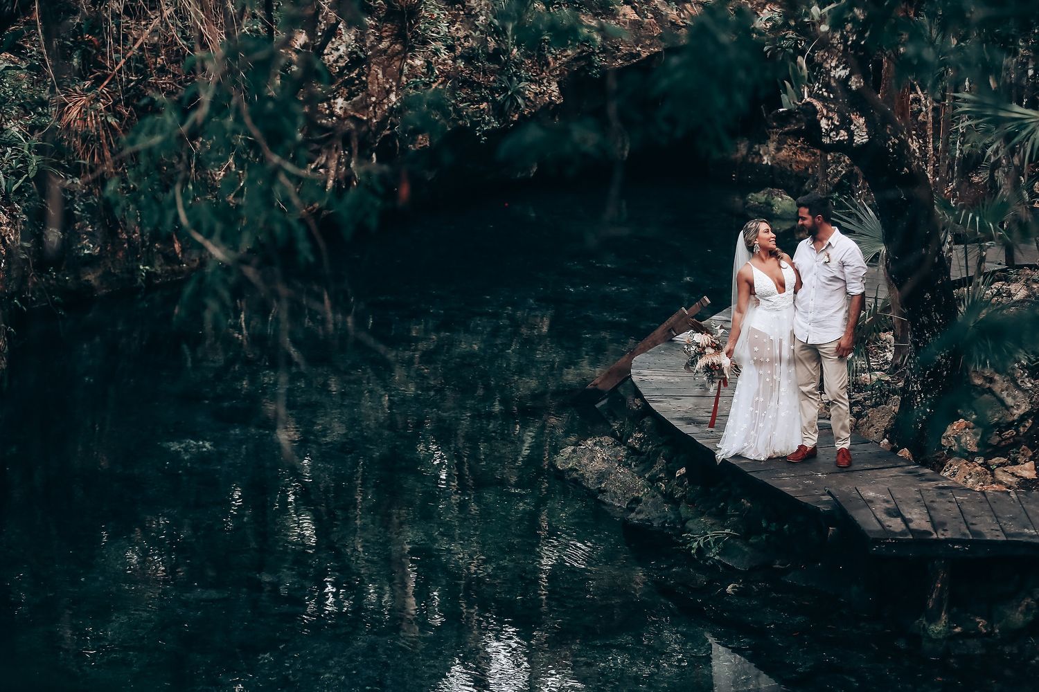 A couple shares an intimate moment on a rock ledge overlooking dark cenote waters.