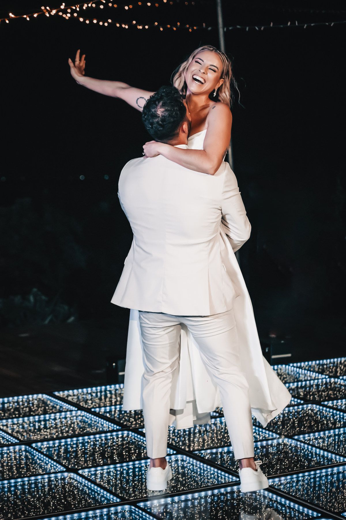 Joyful bride being lifted by groom on illuminated dance floor during outdoor evening wedding reception.